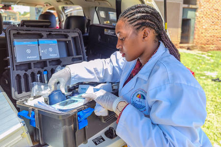 A woman wearing a lab coat and latex gloves reaches into a photometer (mobile water testing kit) set up in the trunk of a van.
