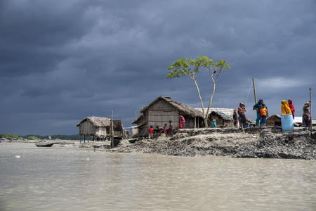 Image is of a village sitting in sand, on the edge of brown water. Small, thatch homes line shore. People wearing bright clothing are pictured on the sand. Dark storm clouds can be seen over the village.