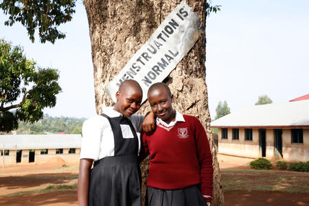 Två gladaflickor står på sin skolgård framför en banderoll med texten "Mens är normalt".