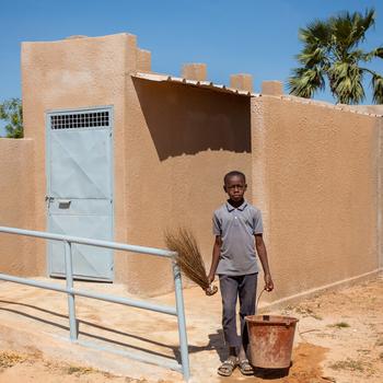 11-year-old Zie stands with a brush and bucket next to his school toilet block in Banfora, Burkina Faso.