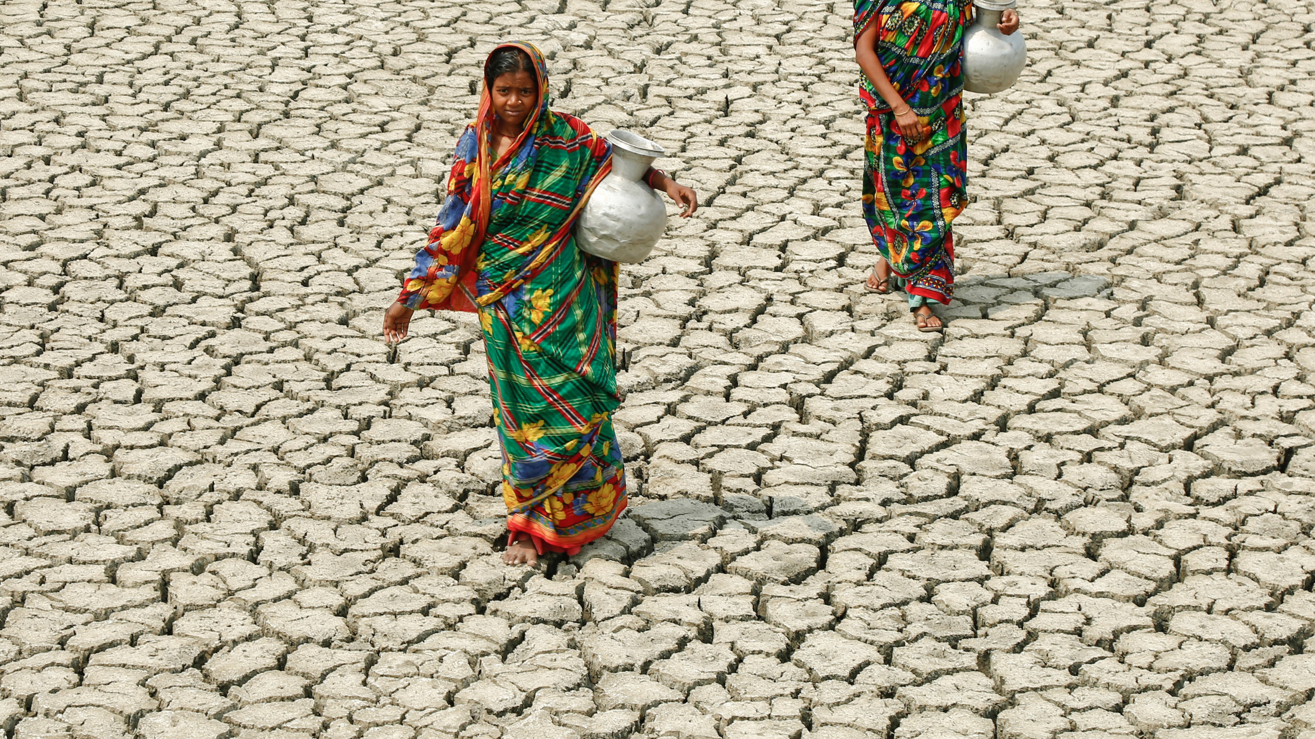 Two women, holding water jugs, walking across extremely dry, cracked earth. 